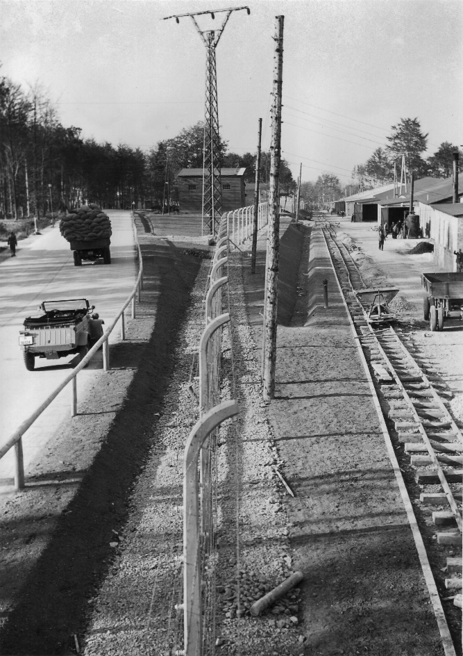 The picture was taken in portrait and from an elevated position. In the centre is the fence that extends into the background of the photo. To the left of the barbed wire fence is a road on which lorries drive. To the right of the fence you can see a factory site on which railway tracks run.