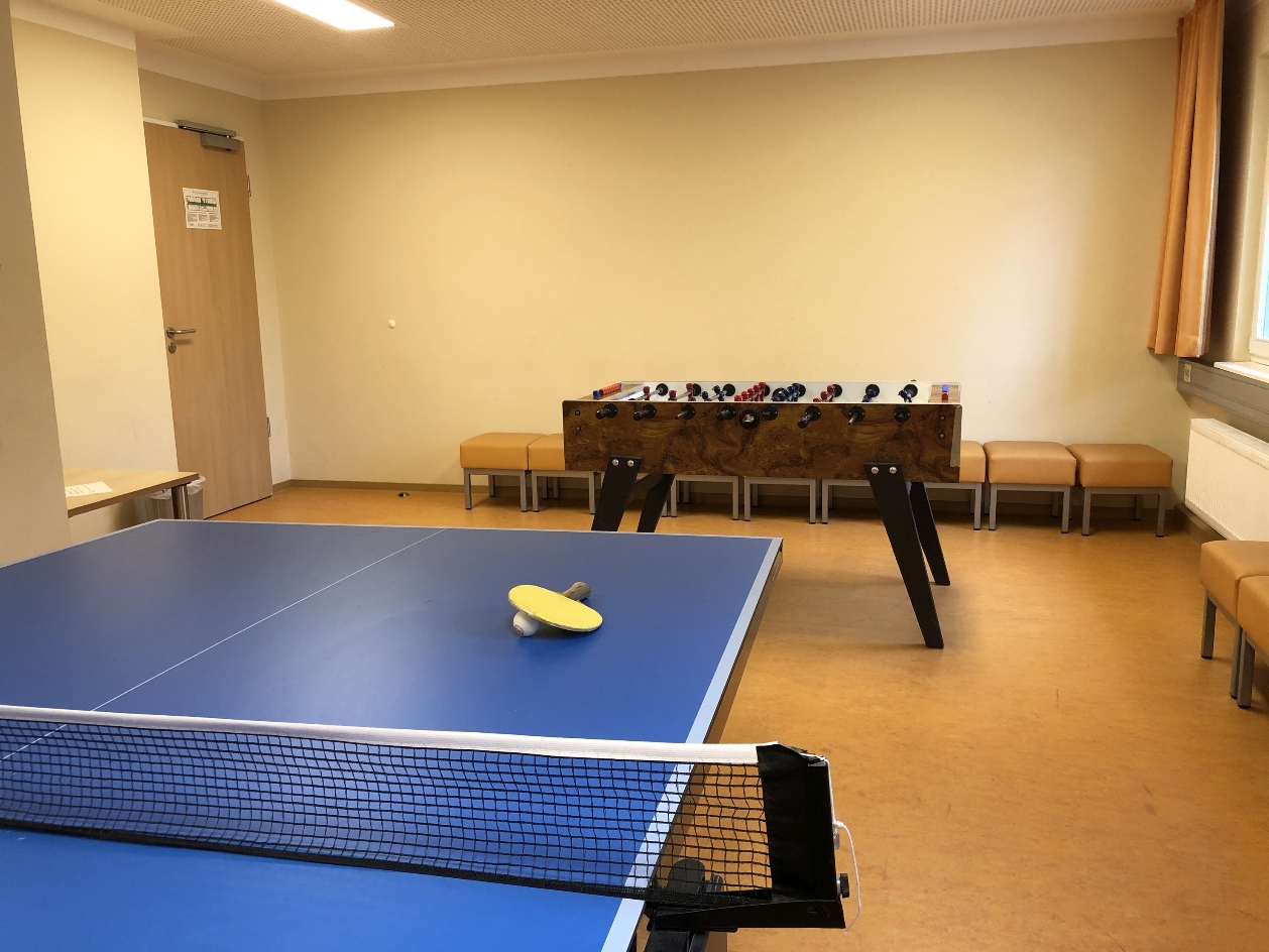 View into a recreation room of the International Youth Meeting Center. In the foreground is a table tennis table with a racket and a ball. In the background you can see a foosball table.