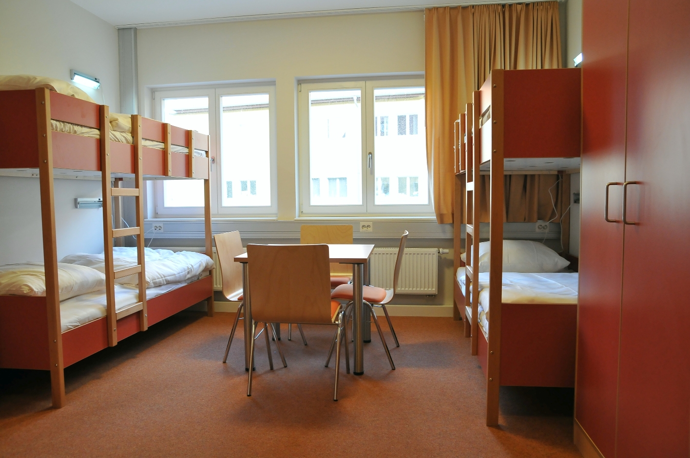 A guest room in the International Youth Meeting Center. To the left and right of the wall are two double bunk beds, in the middle a table with four chairs, behind them are several windows.