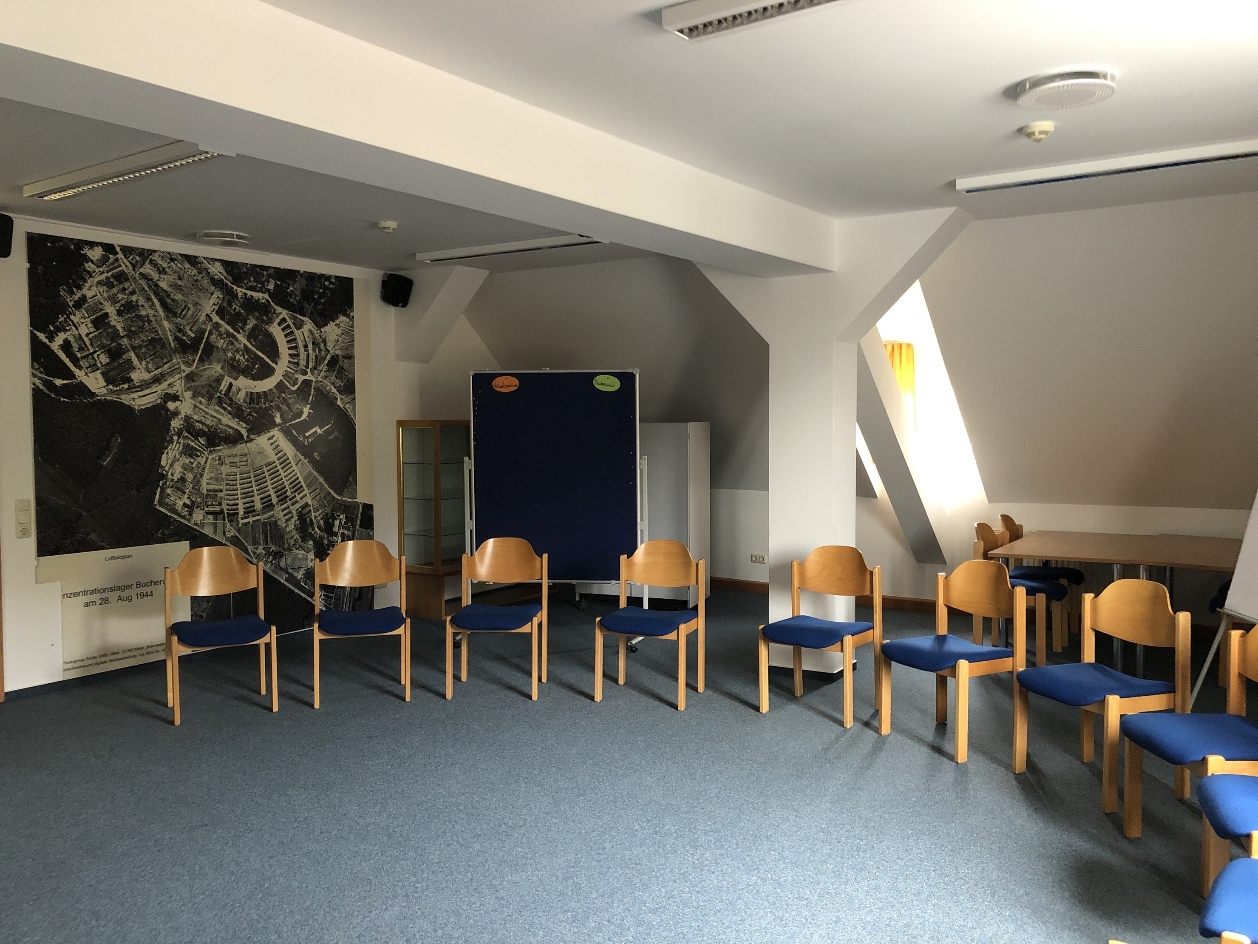 View into a seminar room of the International Youth Meeting Center. There are chairs in a semicircle. On the wall is an aerial photograph of the former Buchenwald concentration camp. Next to it is a pinboard.