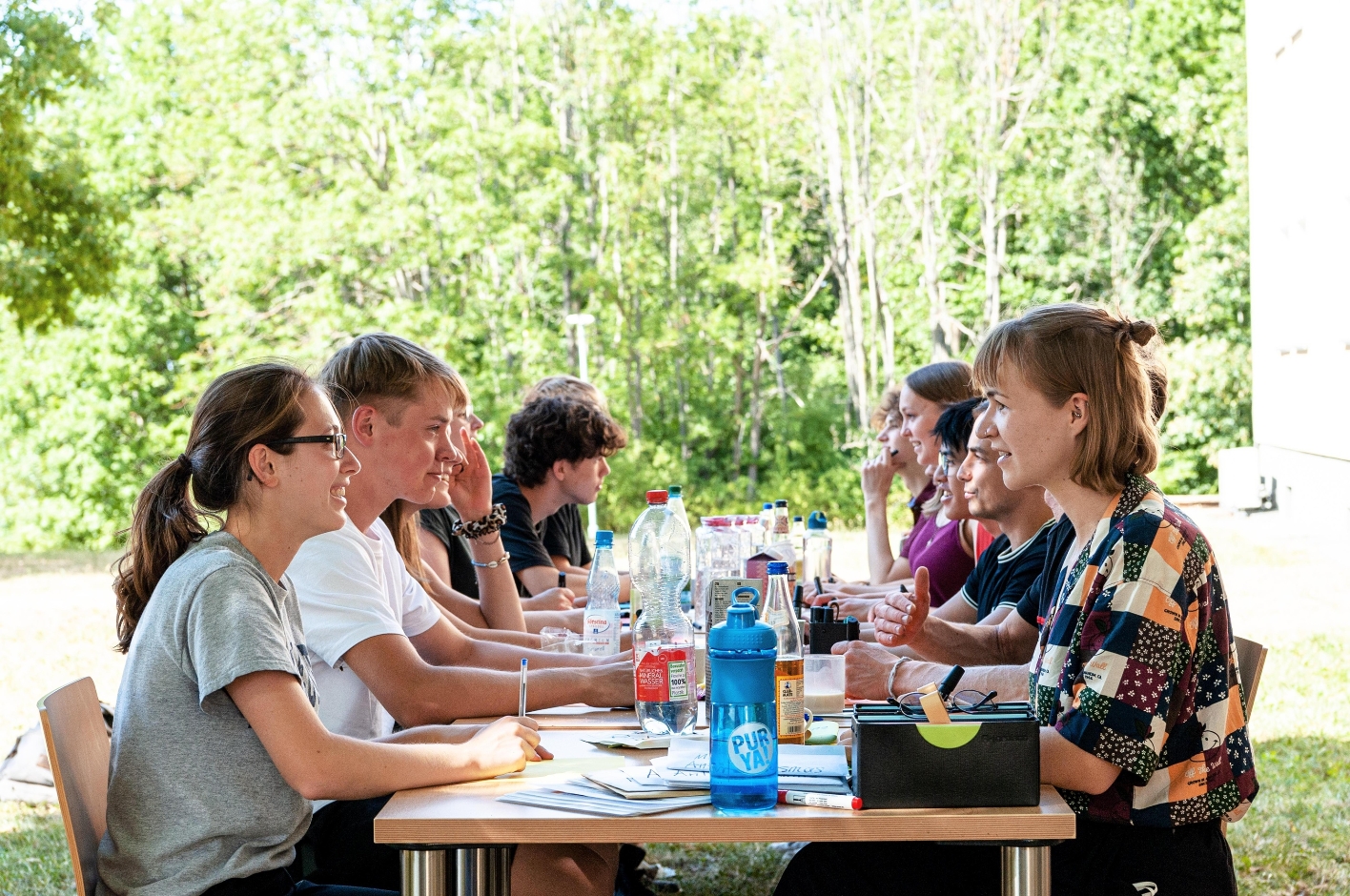 Zu sehen sind mehrere Jugendliche an einem Tisch in der Nähe der Jugendbegegnungsstätte, die angeregt miteinander diskutieren. Neben Getränken ist Lehrmaterialauf den Tischen verstreut.