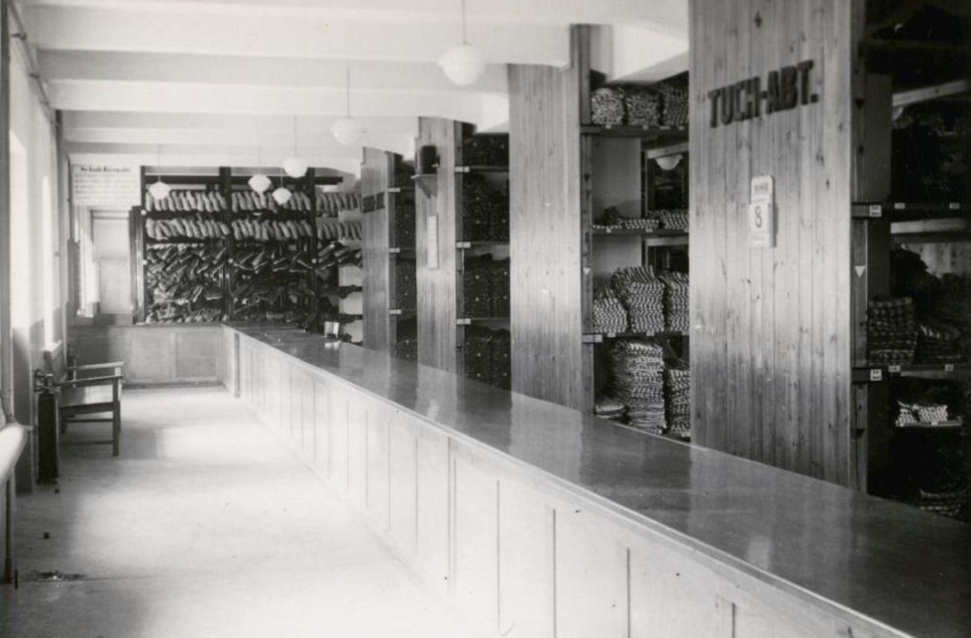 The picture shows a longer room divided into two areas by a long counter. On the right you can see a corridor and windows on the wall. On the left behind the counter, four large shelves can be seen, which are stocked with clothes and hangers. At the very back of the room, a shelf with wooden shoes can be seen.