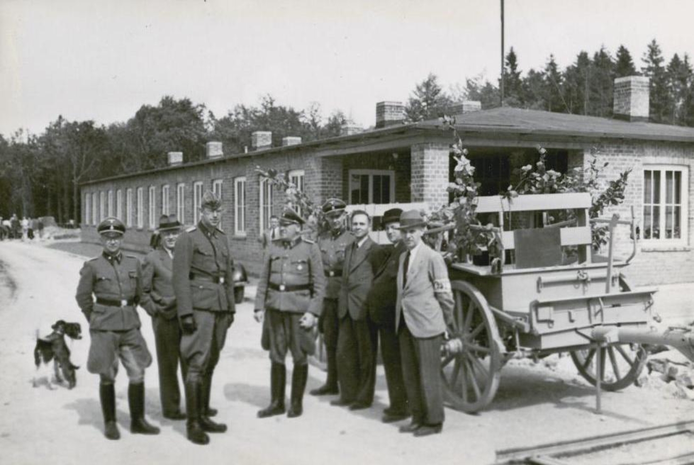 Lagerkommandant Pister (Mitte) und SS-Männer mit Betriebsleitern der Rüstungsfabrik. Die Männer stehen vor einem Pferdegespann. Im Hintergrund ein Bungalow.