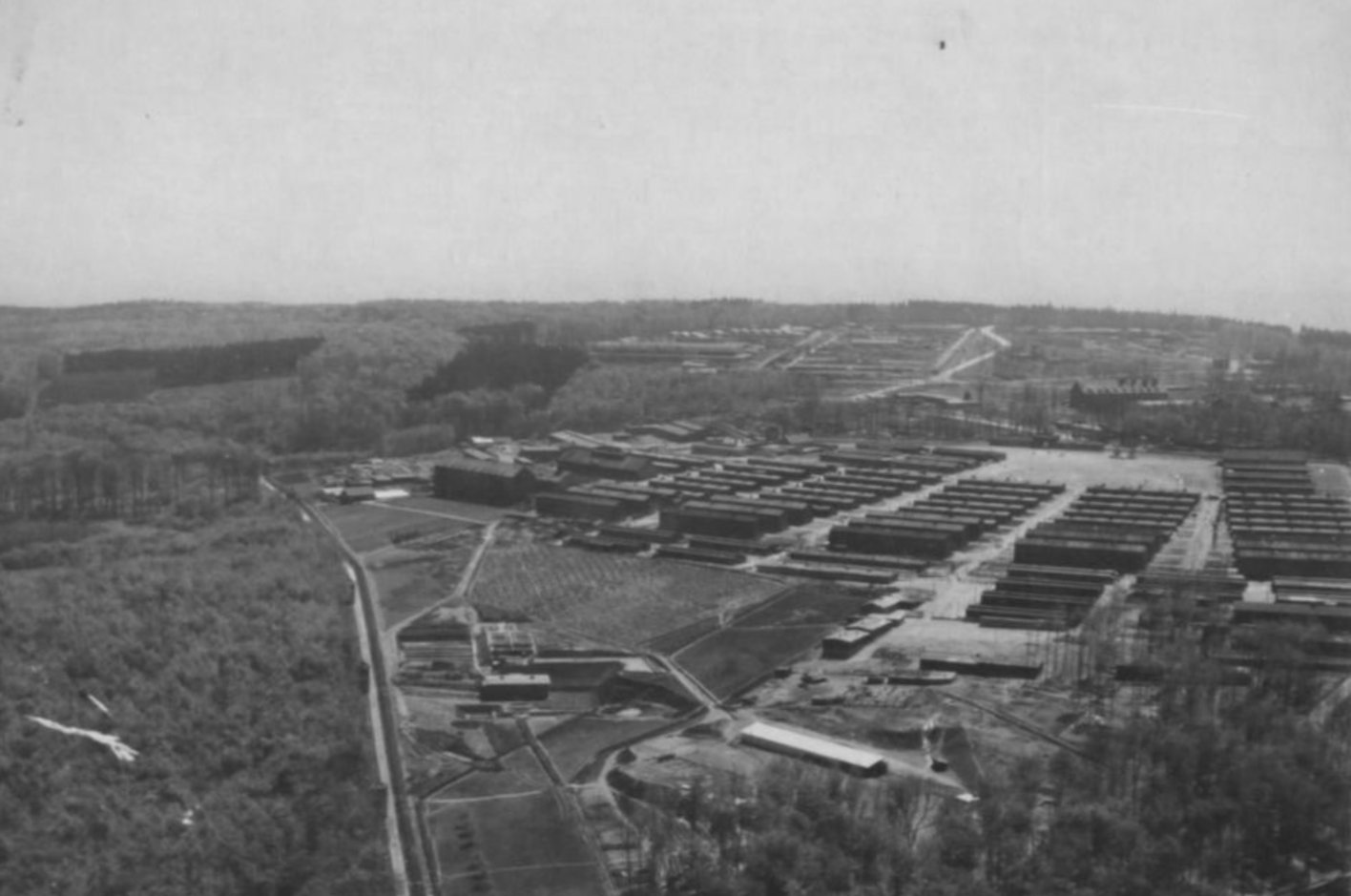 Low-level aerial view of the concentration camp from the northwest. In the center of the picture the prisoners' camp with rows of barracks, in the background the partly destroyed Gustloff factories. Surrounded by forest.
