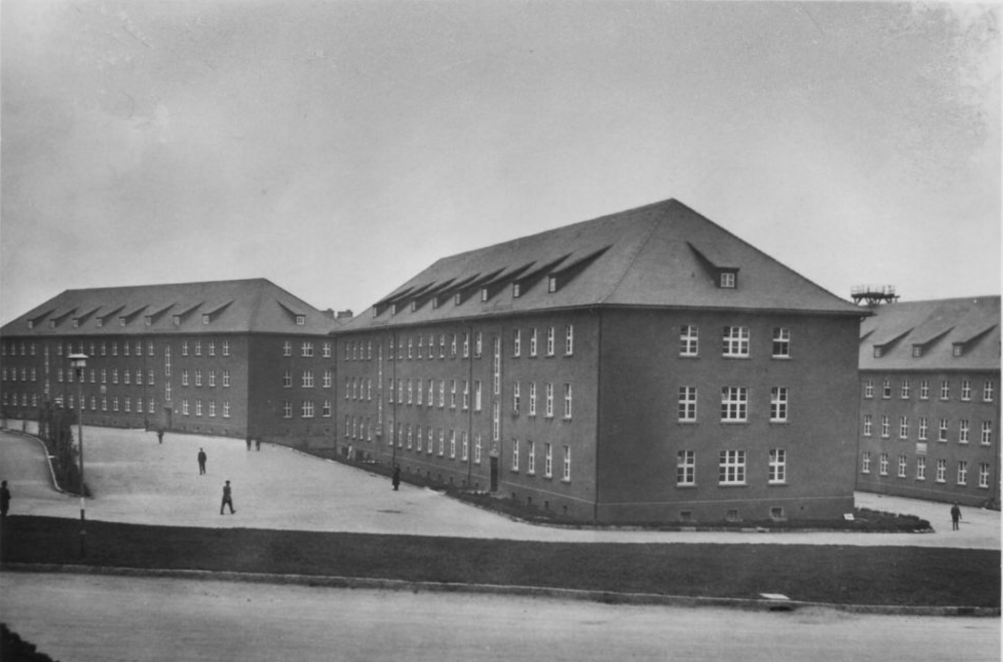 In the picture you can see a total of 3 barracks buildings built in the same style. All three are elongated and have three floors. In front of them a few people.