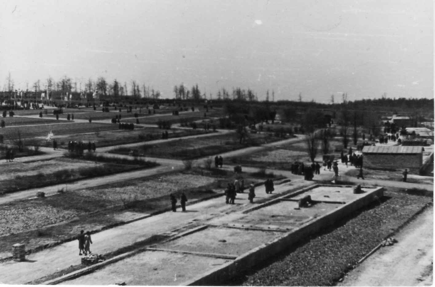  View from the chamber building to the southwest, in front the foundation of stone barrack No. 13. The two barrack fields in the center of the picture indicate the location of the so-called isolator (barracks No. 19 and No. 20), two barracks separated from the rest of the camp and surrounded by a high barbed wire fence. The barracks are only visible through their foundations. Numerous people walk on paths across the grounds.