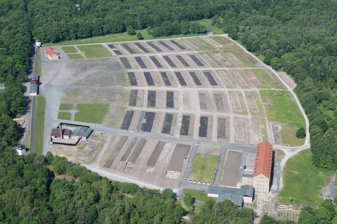 Blick aus der Luft auf das ehemalige Häftlingslager. Dunkle Flächen markieren die Grundrisse der Baracken. Wenige Gebäude stehen noch. Rundherum Wald.