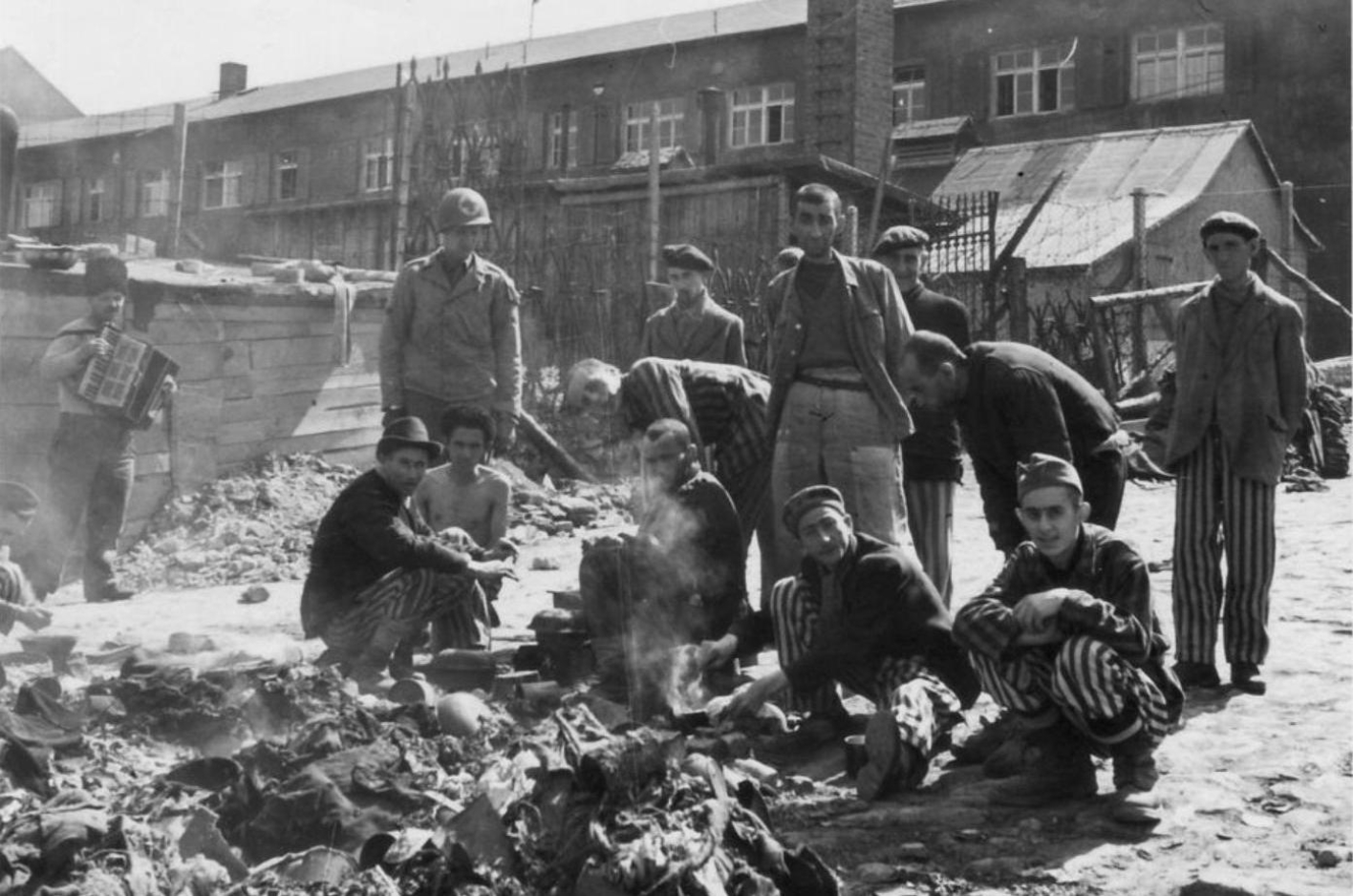 Surviving prisoners of the Small Camp cook a meal among rubble and debris. Five squat on the floor, five more stand. On the left stands a U.S. Army medic. Also, a man with an accordion leans against a wall.