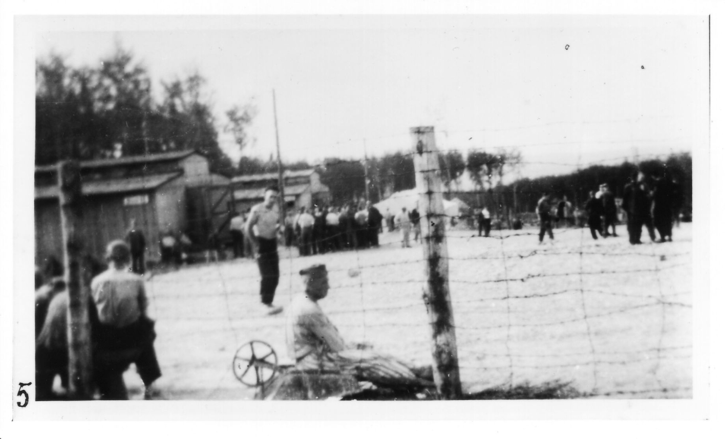 Häftlinge im Kleinen Lager, durch einen groben Stacheldrahtzaun fotografiert. Links die Pferdestallbaracken 62 und 63. Mit dem Rücken zum Zaun sitzen einige Häftlinge. Im Hintergrund ist das im Mai 1944 errichtete Zeltlager zu erkennen. Im Hintergrund sind Gruppenansammlungen von Häftlingen zu sehen.