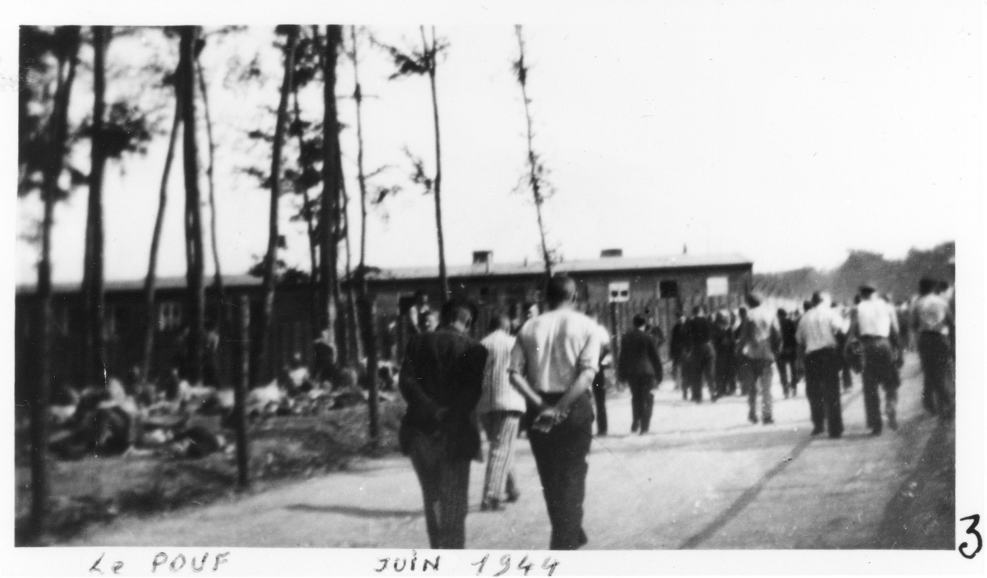 Häftlinge laufen auf der Lagerstraße in Richtung einer Baracke, die im Hintergrund zu sehen ist. Die Häftlinge laufen in kleinen Gruppen und machen den Eindruck als würden sie miteinander reden.