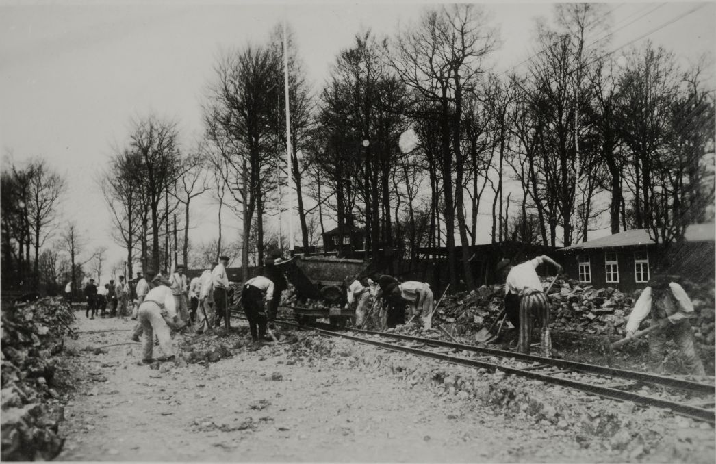 The picture shows people in prisoners' clothing shovelling stones and gravel along tracks. The camp gate can be seen through the treetops in the background.