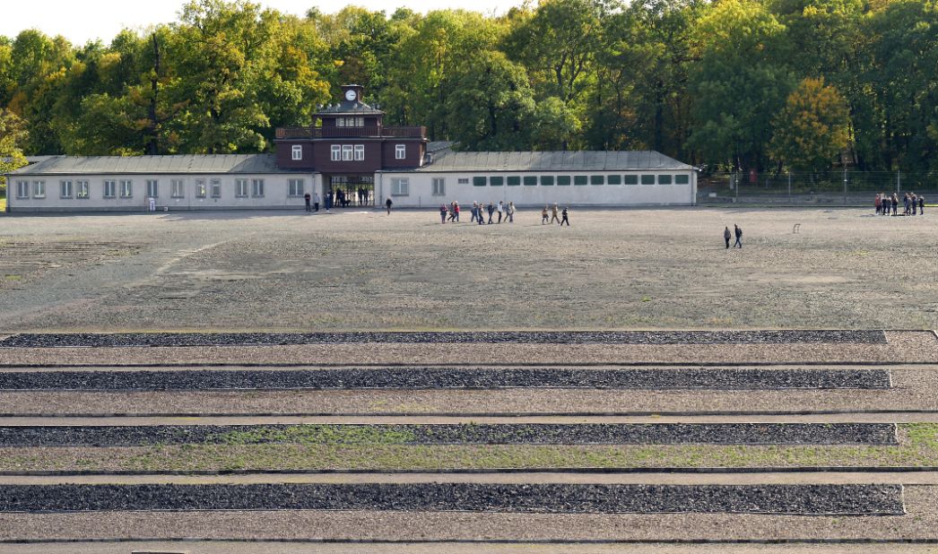 The former roll call area can be seen as a large open area in front of the camp gate, which can be seen in the background. In the foreground of the picture, the outlines of the former barracks of the camp are made visible on the ground by fields of cinder stones, which stand out darkly against the rest of the gravel.