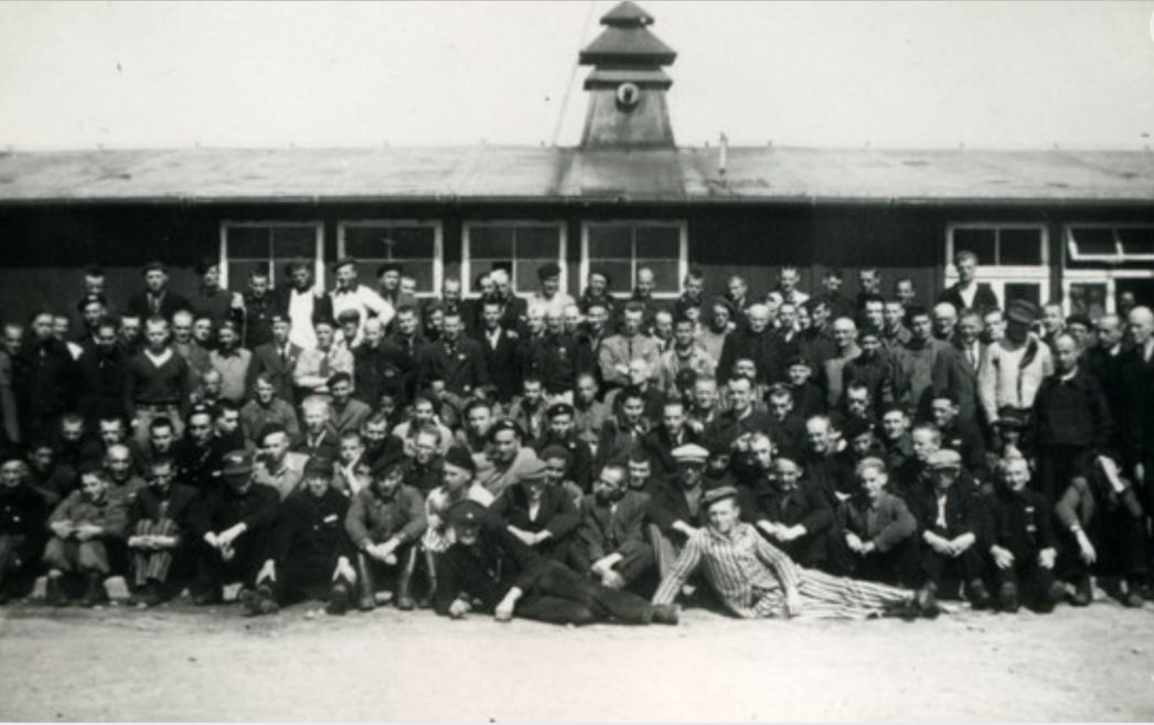 Group photo of liberated Dutch prisoners in front of a wooden barrack. Occasionally, some still wear prisoner clothing.