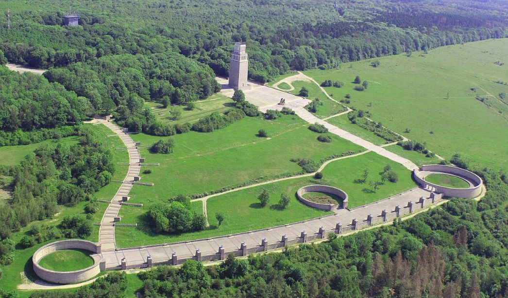 Aerial view of the memorial site. At the top left, the path begins downhill. Further down, the balcony-like Street of Nations stretches from one ring grave to the next. Halfway down, another ring grave. From there uphill the path leads to the bell tower.