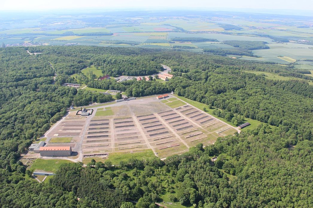 The area of the memorial photographed from the air. On the site you can see individual buildings and open spaces. The area is surrounded by trees. The foundations of the barracks are still visible. 