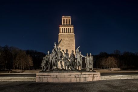 The group of figures at the Buchenwald Memorial in front of the memorial's bell tower at night. Both the group of figures and the bell tower are illuminated.