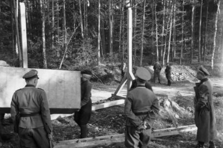Im Vordergrund stehen drei Männer in SS-Uniformen, vor ihnen tragen andere hölzerne Einzelteile zu zwei aufgestellten Pfosten, die bereits als Galgen im Aufbau zu erkennen sind.