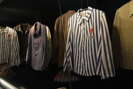 Six different tops of prisoners' clothing: Three of them blue and white striped, two olive and one dark blue. On the three in front there are red triangles on the top at chest level, with a number underneath.
