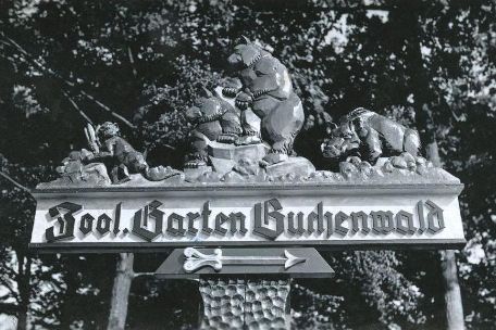 A hand-carved signpost with the inscription "Zool. Garten Buchenwald" (Zoological Garden), with animal figures above the lettering: a monkey, two bears and a wild boar.