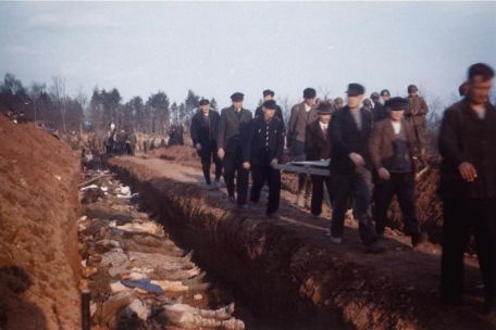 Residents of Nordhausen, under the supervision of American soldiers, bury the dead of the Boelcke Barracks concentration camp in a row grave. On the right side of the trench, some men carry a stretcher with a dead body past. The trench is filled tightly with corpses.