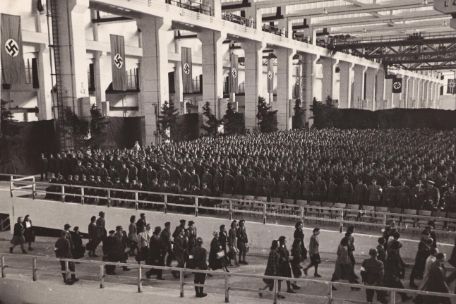 The rocket assembly hall F 1 in Peenemünde, which is characterized by its size and height. In the foreground, people are moving into the hall. Behind it, people can be seen lining up for roll call. Near the side wall of the hall are several swastika banners.
