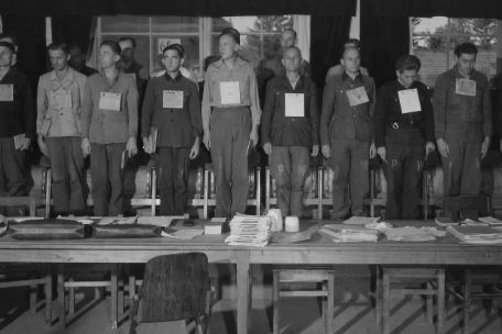 A photo of the defendants standing on their chairs. They all have a sign hanging around their necks with their name and charges listed.