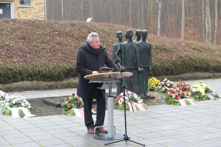 Prof. Dr. Wagner bei einer Ansprache auf dem Gedenkplatz vor dem Krematorium. Er ist dunkel gekleidet und steht an einem schmalen Rednerpult. Im Hintergrund sind Blumenkränze, die um die Gedenktafel und Figurengruppe des Gedenkplatzes arrangiert wurden.