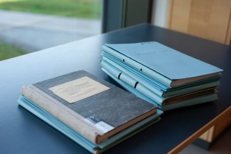 Auf einem Tisch liegen ein altes Notizbuch und ein Stapel blauer Mappen.