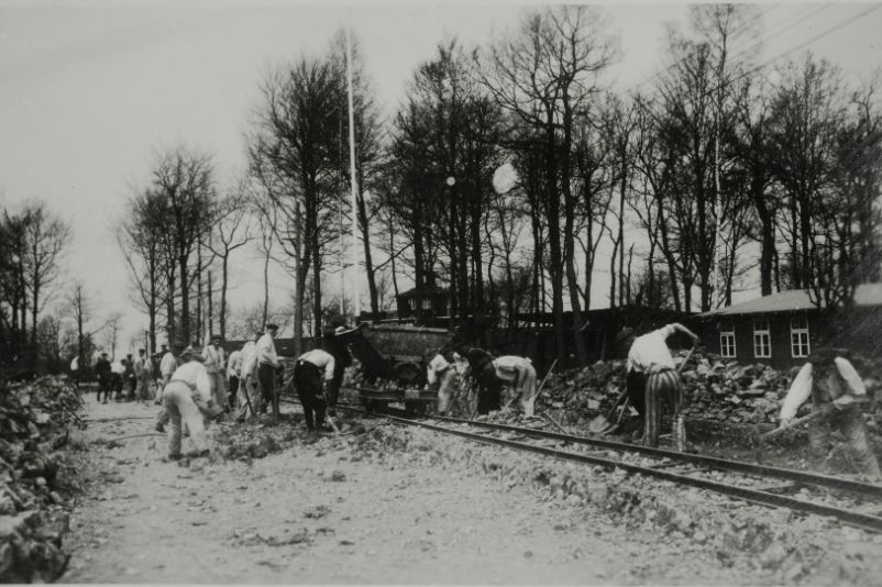 The picture shows people in prisoners' clothing shovelling stones and gravel along tracks. The camp gate can be seen through the treetops in the background.