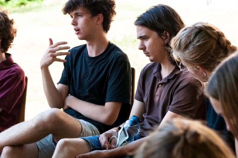 Part of a group of young people can be seen exchanging opinions. They are sitting outside on a meadow in a circle of chairs and discussing.