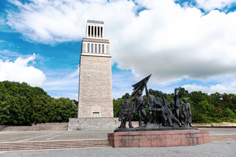 In the foreground is a larger-than-life bronze figurine group staging prisoners of the concentration camp after their self-liberation. In the background rises the bell tower of the memorial complex, built of light stone.