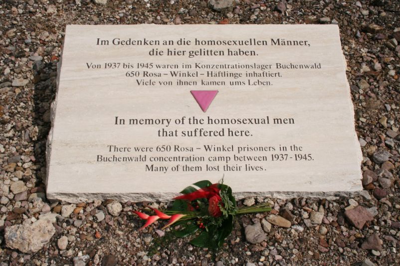 Weißer Gedenkstein mit dem rosa Winkelzeichen für Homosexuelle Häftlinge im Zentrum. Gravur: „Im Gedenken an die homosexuellen Männer, die hier gelitten haben. Von 1937 bis 1945 waren im Konzentrationslager Buchenwald 650 Rosa-Winkel-Häftlinge inhaftiert. Viele von ihnen kamen ums Leben" In Deutsch und Englisch.