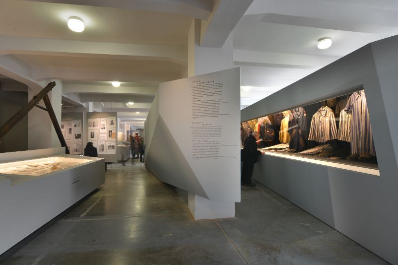 View into the exhibition. On the right, abstractly shaped showcases display items of clothing, and on the left, an aisle with more showcases displaying documents and photographs. 