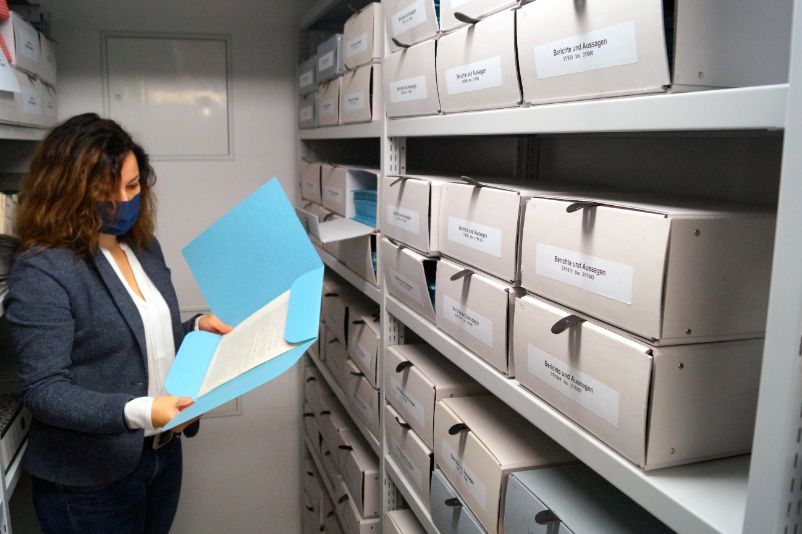 Eine Mitarbeiterin steht neben einem Regal voller Kisten, die mit "Berichte und Aussagen" beschriftet sind. Sie hält eine blaue Mappe in der Hand.