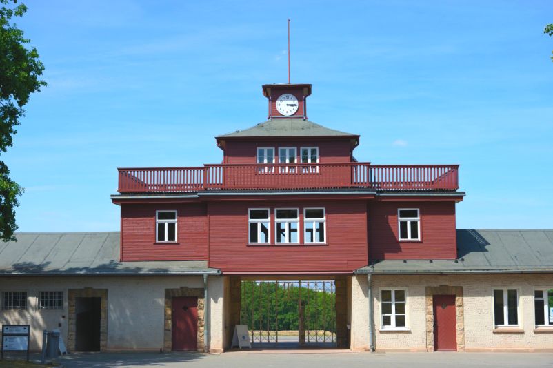 View of the gate building from the former Haeftlingslager site. The gate building has two wings. The left one is characterized by blinded cell windows. The right one has larger windows. In the center of the building there is the main wooden watch tower with a clock on another small structure on the roof.