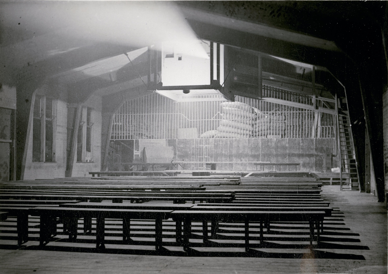 Zusehen ist eine Leere Baracke in der in der Mitte Bänke aufgestellt wurden. Das Bild wurde von der Leinwand aus gemacht. Die Bänke sind der Leinwand zugewandt. An der Decke hängt in einem Großen Kasten ein Projektor aus dem ein Lichtkegel nach vorne scheint.