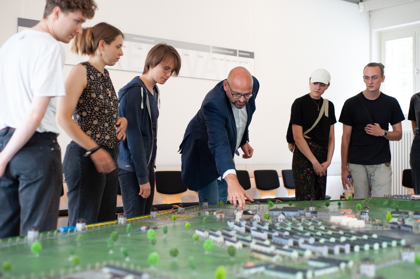 An employee of the memorial points to a model of the concentration camp filling the seminar room. Around the model stand young seminar participants, 5 of whom can be seen in the frame.
