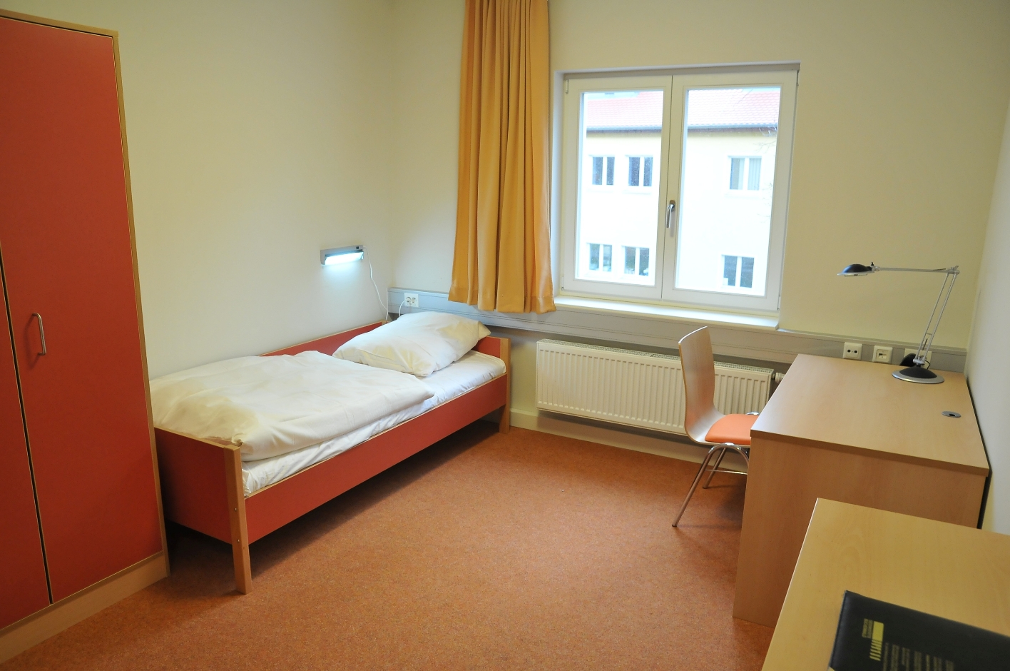 Ein Gästezimmer in der Internationalen Bildungsstätte der Gedenkstätte Buchenwald. Links steht ein Schrank, daneben ein Einzelbett, dann kommt ein Fenster. An der gegenüberliegenden Wand steht ein Schreibtisch mit einem Stuhl und ein kleinerer Schrank.