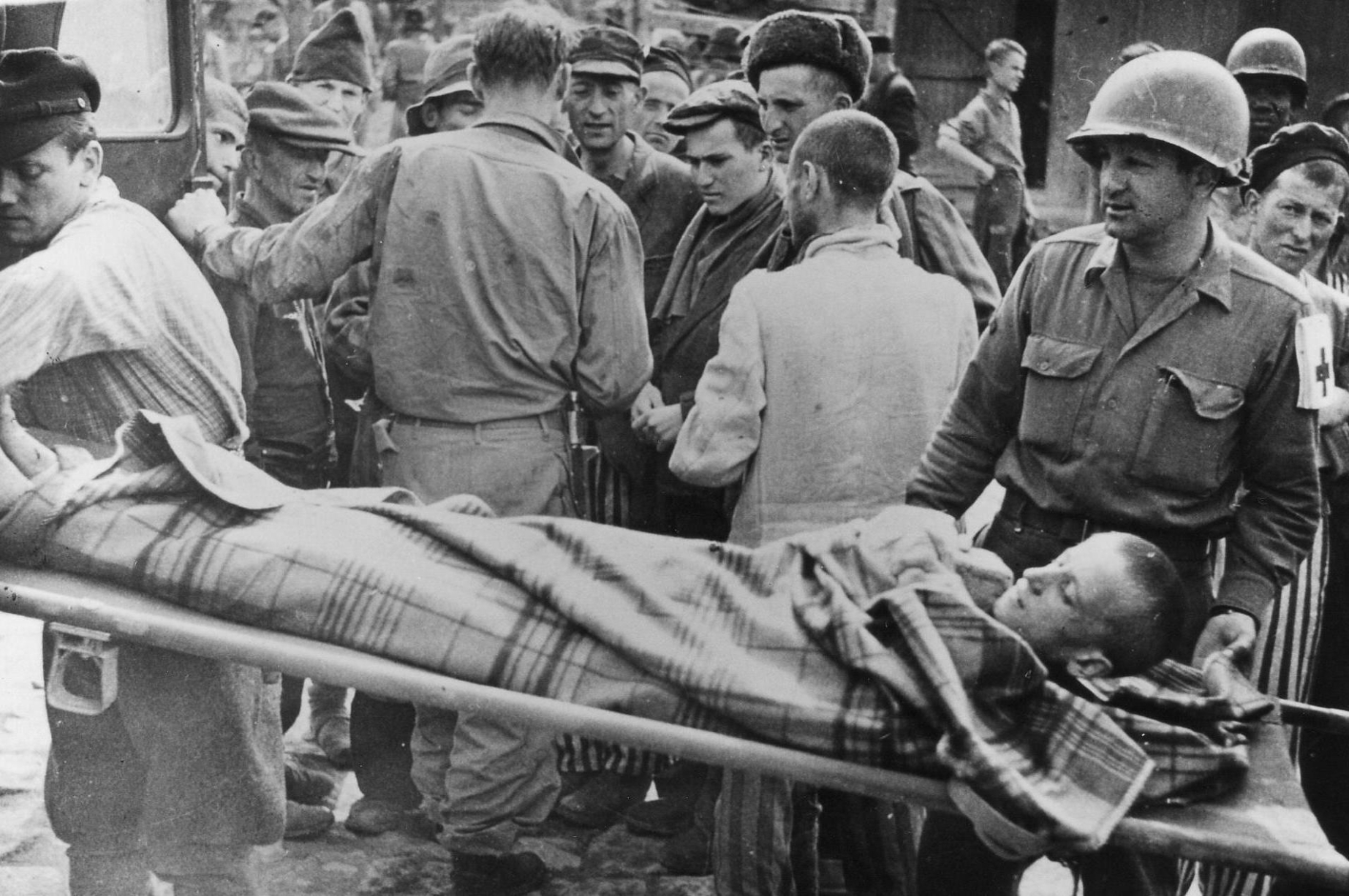 Ein auf einer Bahre liegender, in eine Decke eingehüllter Mann wird in einen Transporter geladen. Ein Soldat mit Sanitäter-Zeichen führt die Trage. Im Hintegrund stehen Angehörige der U.S. Armee sowie befreitete Häftlinge.