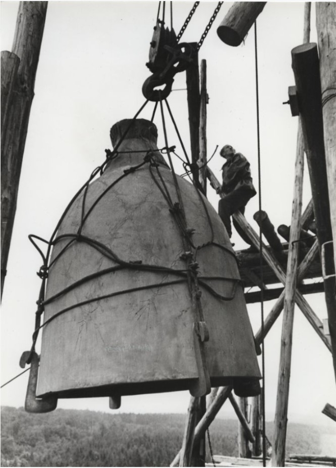 Ein Arbeiter sitzt auf dem eingerüsteten Glockenturm und beobachtet das Heraufziehen der Buchenwaldglocke. Die Glocke ist erheblich größer als der Arbeiter.