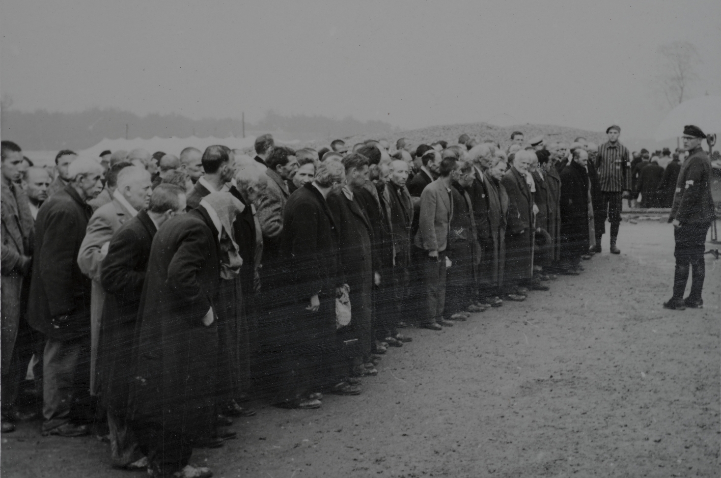 Men dressed in civilian clothes are lined up. They look visibly worried. A man in SS uniform stands in front of the men who are getting ready and speaks to them.