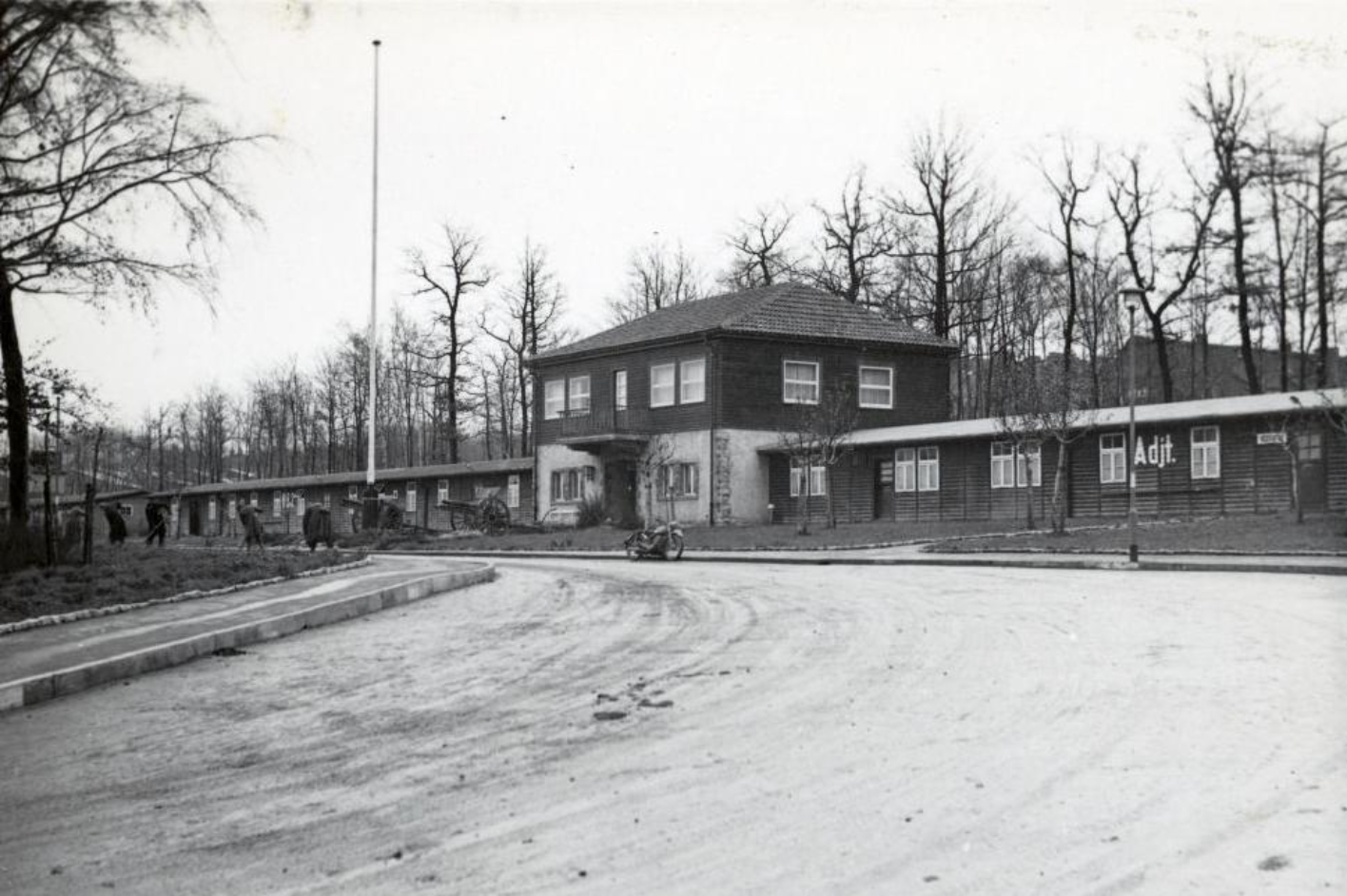 View from the camp gate across Carachoweg to the building of the camp commandant's office and adjutant's office. The building consists of two flat-built wings and a smaller central section with two floors. 