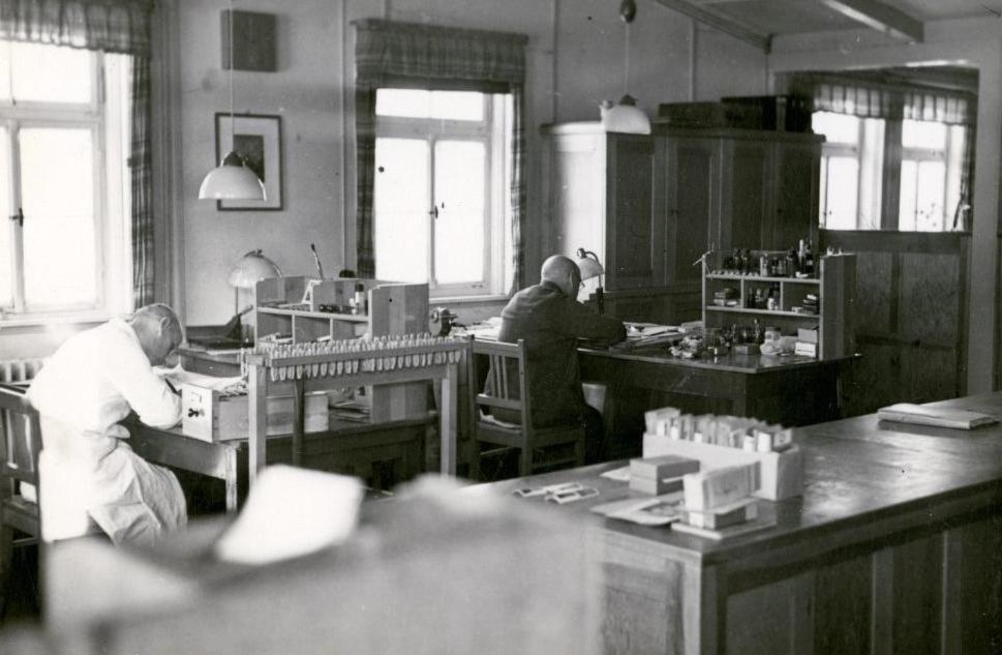 Two prisoners at archival work in the office of the recognition service photographed diagonally from behind. On the right, presumably the prisoner Karl Siebeneichler dressed in white. On the left, another prisoner dressed in black.