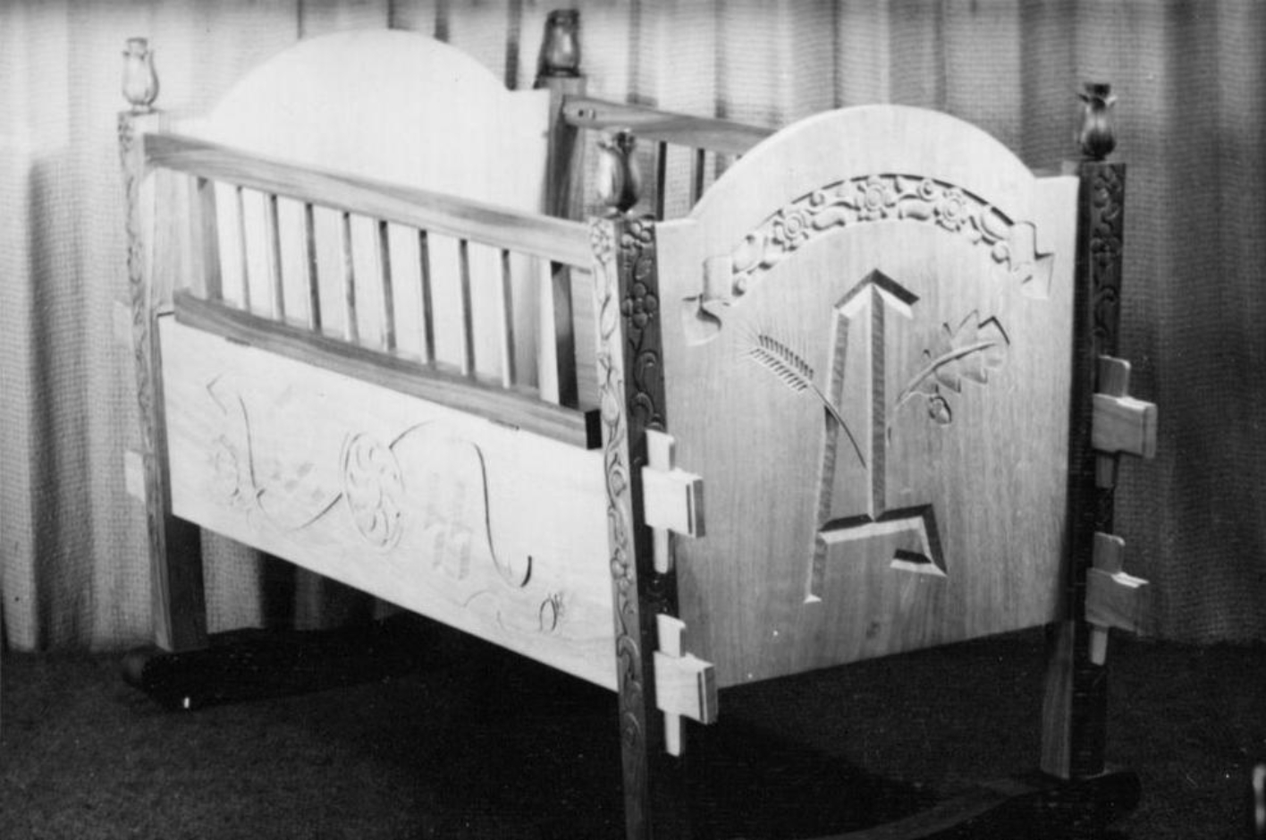 A child's cradle decorated with hand carvings. The cradle is set in scene with view fraom the right on the decorated front and side walls.