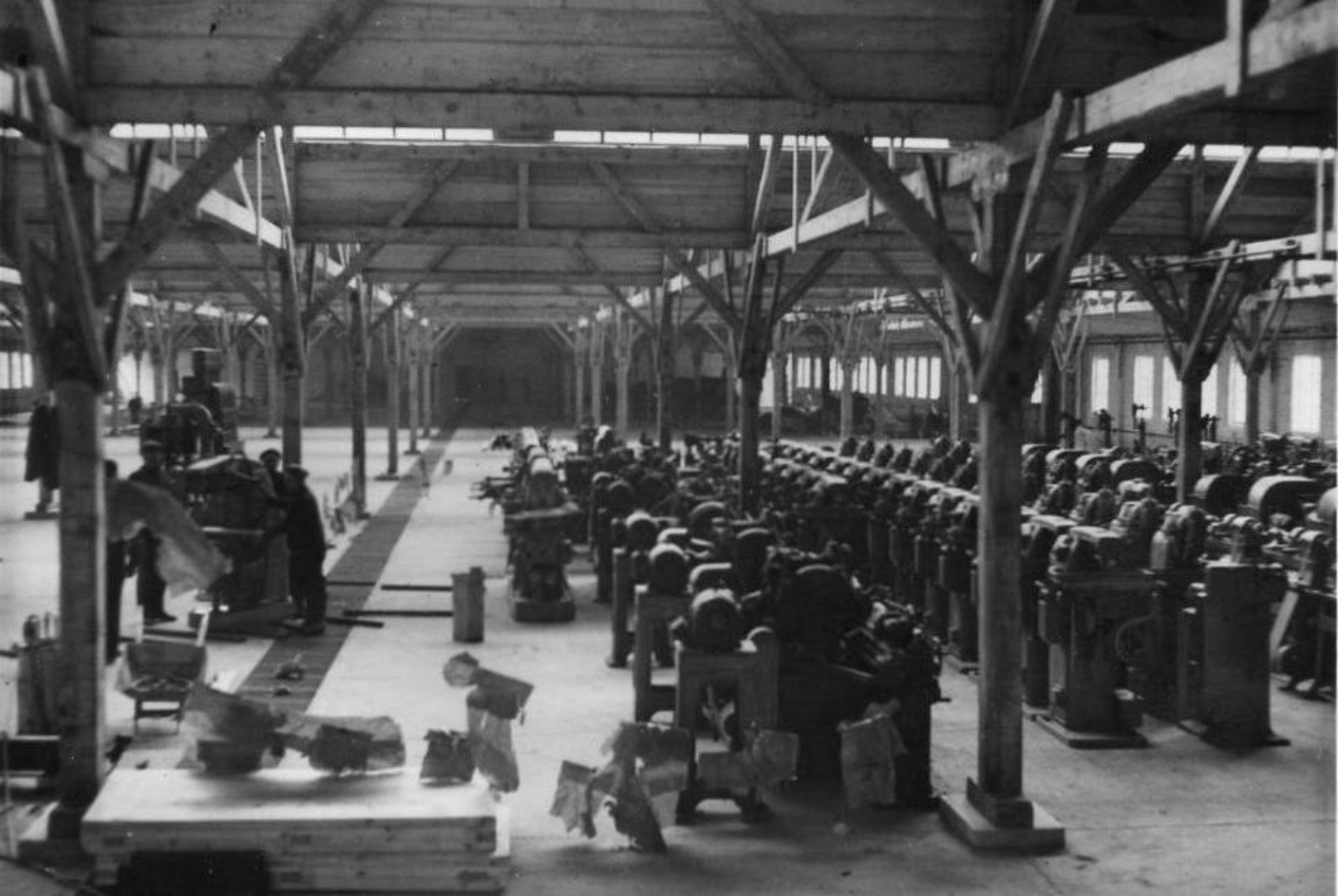 Interior view of the workshop hall at Gustloff Plant II. The hall is partially filled with workbenches arranged in rows. Four workers stand at a single machine.