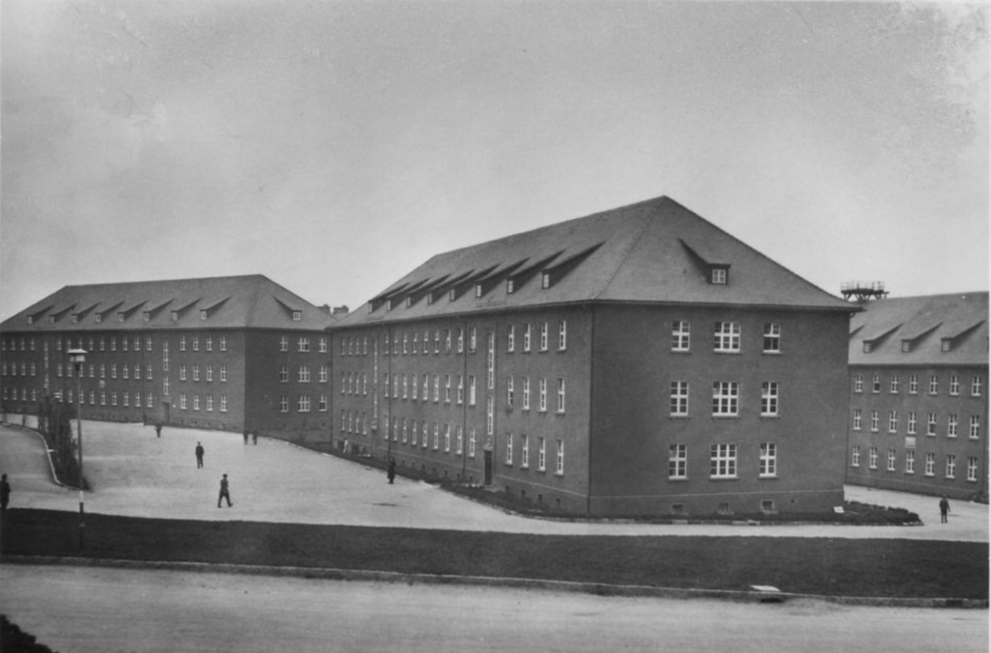 Auf dem Bild sind insgesammt 3 in der gleichen Bauart erichtete Kasernengebäude zu erkennen. Alle drei sind länglich und haben drei Stockwerke. Davor einzelne Menschen.