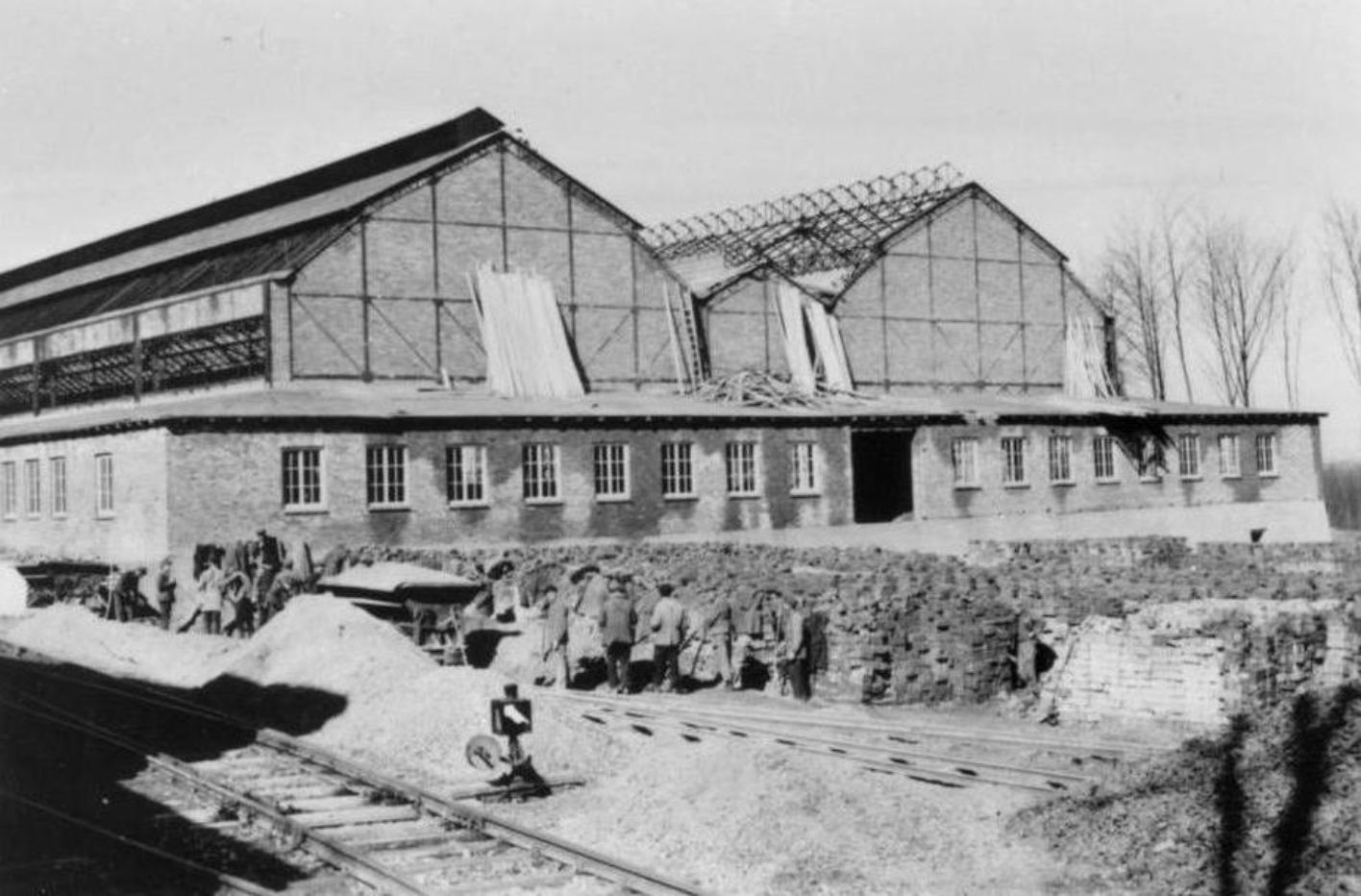 Gleisanschluss der Buchenwald Bahn in den Gustloff-Werken II. Im Hintergrund arbeitende Häftlinge. Vom Werk ist das im Bau befindliche, zweigeteilte Dach einer großen Halle zu sehen. Baumaterial ist an verschiedenen Stellen aufgeschichtet.