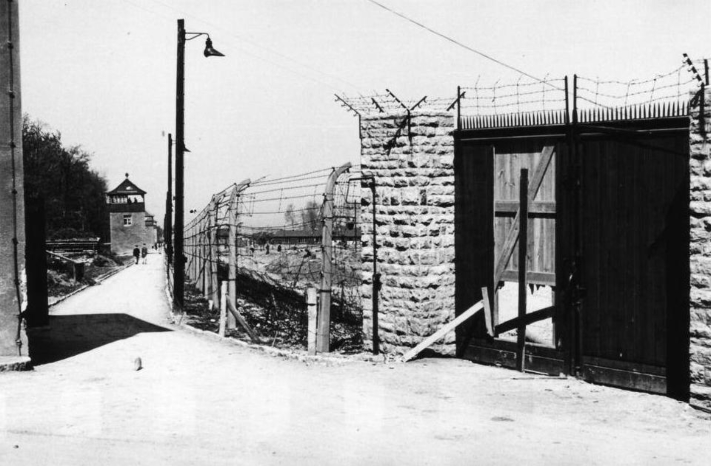 On the left, the post path runs straight along the camp fence. Guard towers can be seen at regular intervals along the path. On the right side of the picture, a gate in the camp fence, provisionally closed with wooden boards, can be seen. The fence consists of high posts that are bent inward at the top, into the camp. Barbed wires are stretched between them. 