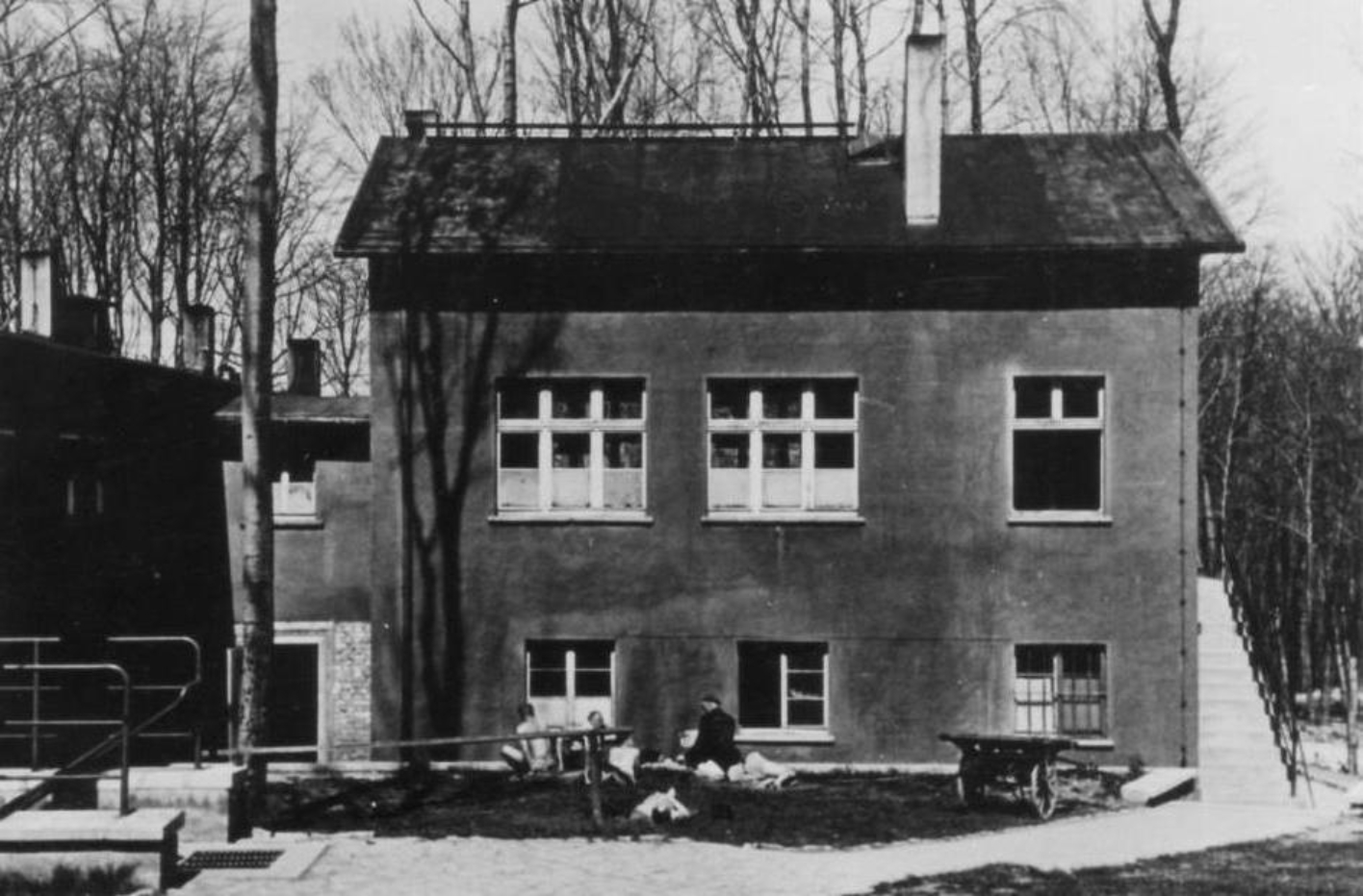 Frontal view of the building that housed Operation Area II of the prisoner infirmary. A few barely clothed prisoners lie and sit in front of the house