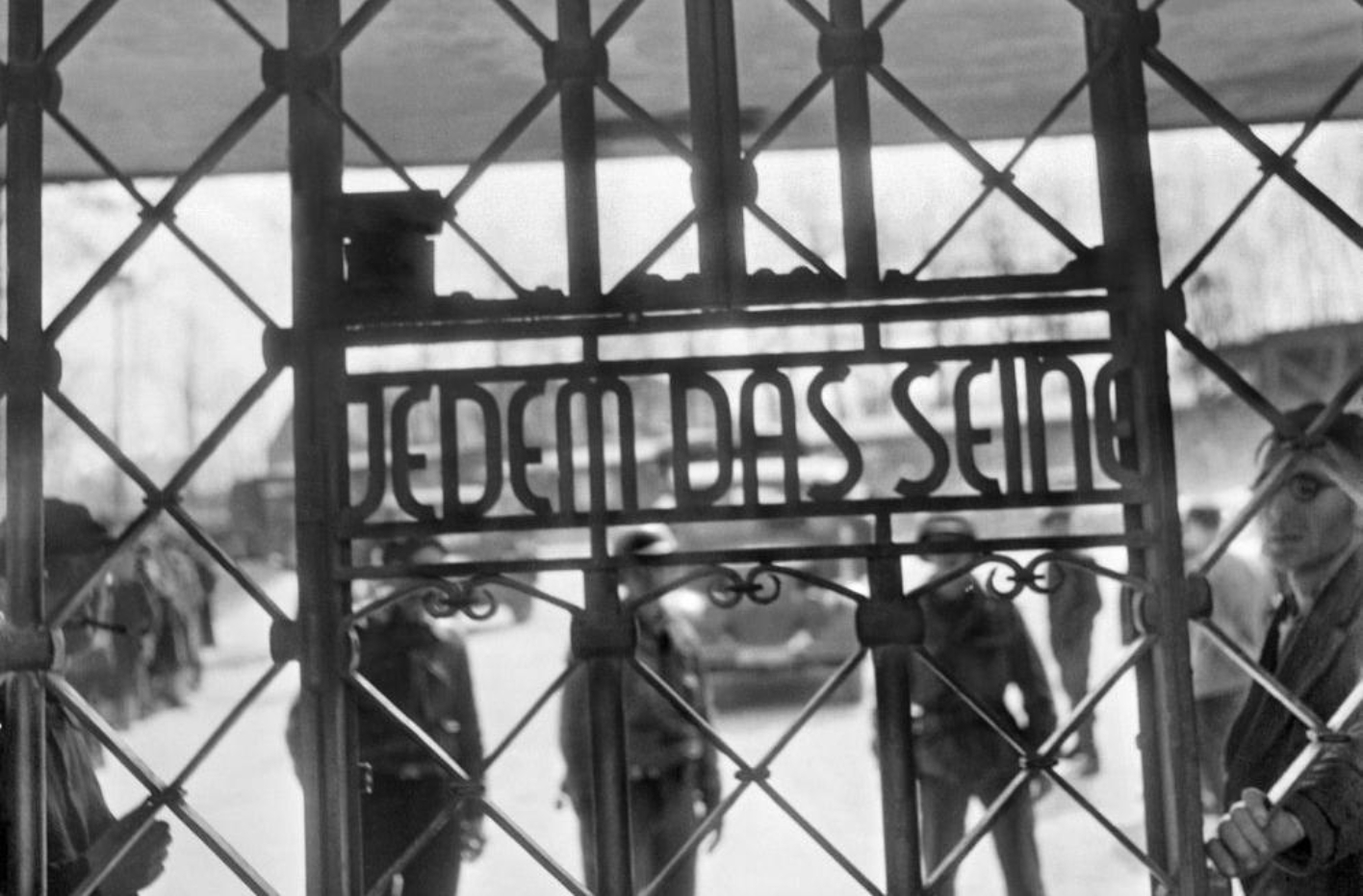 View through the camp gate at the inscription "To each his own". Behind the gate American soldiers and former prisoners.